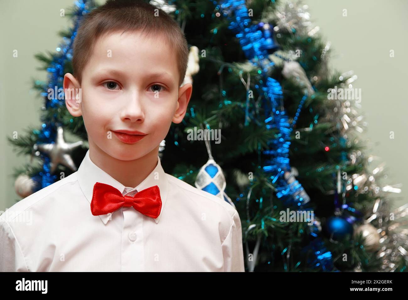 Junge in weißem Hemd und roter Schleife, der neben dem Weihnachtsbaum steht Stockfoto