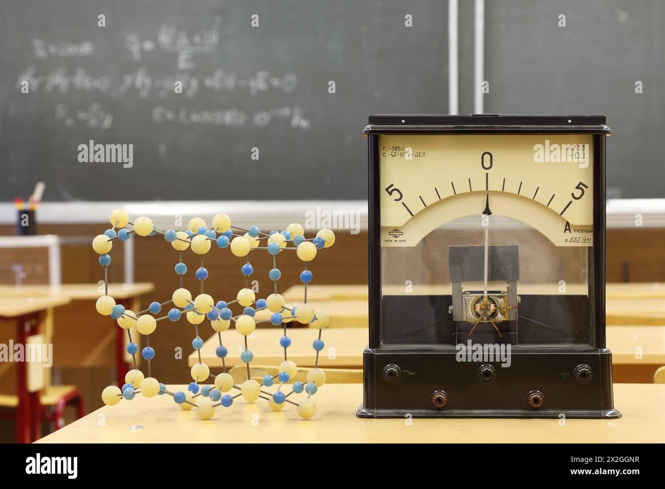 Galvanometer mit nicht realer Zahl 555 und Modell der Atomstruktur auf dem Schreibtisch in leerer Physikschule; Formel auf der Tafel Stockfoto