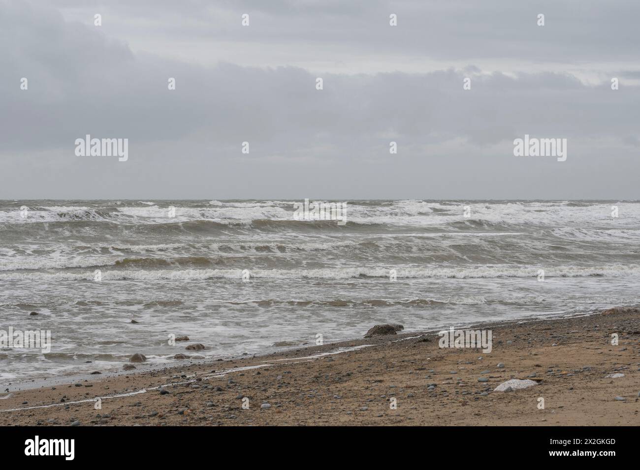 Steiniger Strand unter grauem Himmel mit Wellen im weißen Wasser, stimmungsvolle gedämpfte Meereslandschaft Stockfoto