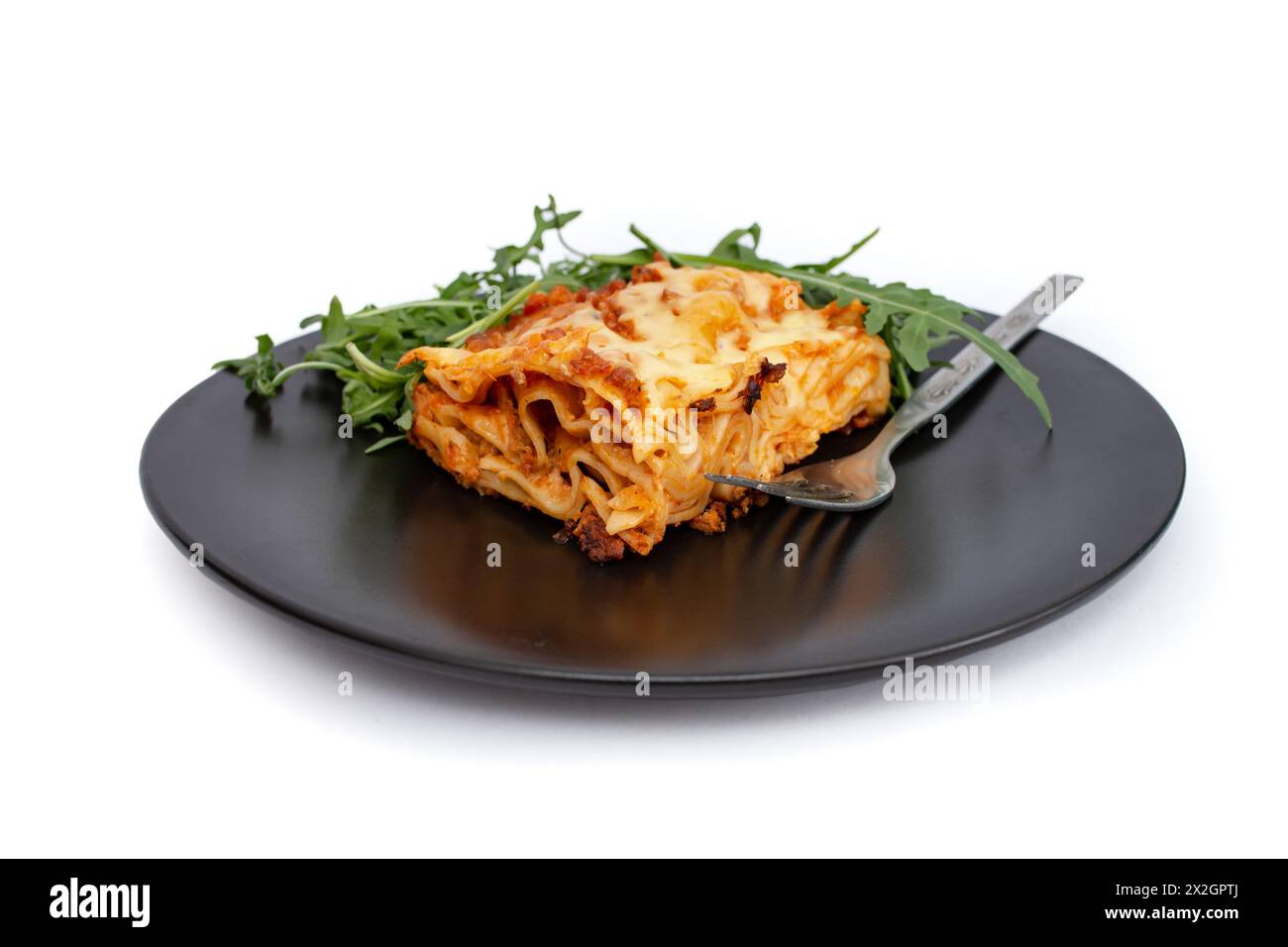 Quadratische Scheibe Lasagne auf einer schwarzen Keramikplatte mit Rucola-Salat, weicher Fokus aus der Nähe isoliert auf weiß Stockfoto