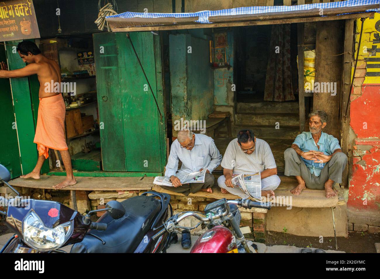 Männer, die Zeitungen lesen, als ein Mann mit Dhoti die Fensterläden seines Ladens in Varanasi, Indien, öffnet Stockfoto