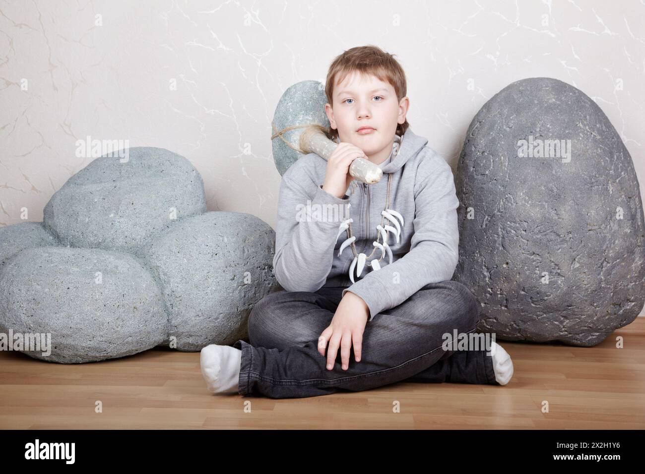 Der Teenager sitzt auf dem Laminatboden mit einer Steinaxt zwischen den Felsen und zeigt den primitiven Mann Stockfoto