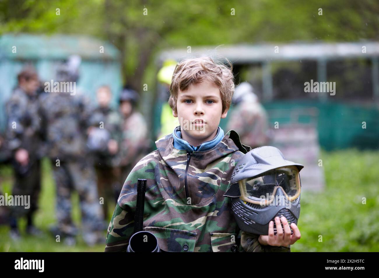 Der Junge in der Tarnung hält ein Paintball-Gewehrlauf in einer Hand und einen Schutzhelm in einer anderen, der mit einer Gruppe von pla auf dem Paintball-Boden steht Stockfoto