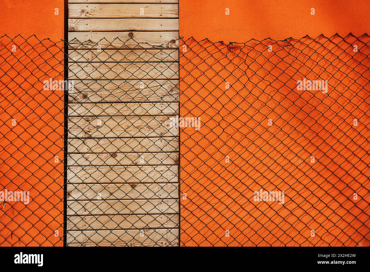 Der Maschendrahtzaun wirft Schatten über die orangefarbene Wand und schließt die Türen mit Holzdielen, Designelement Stockfoto