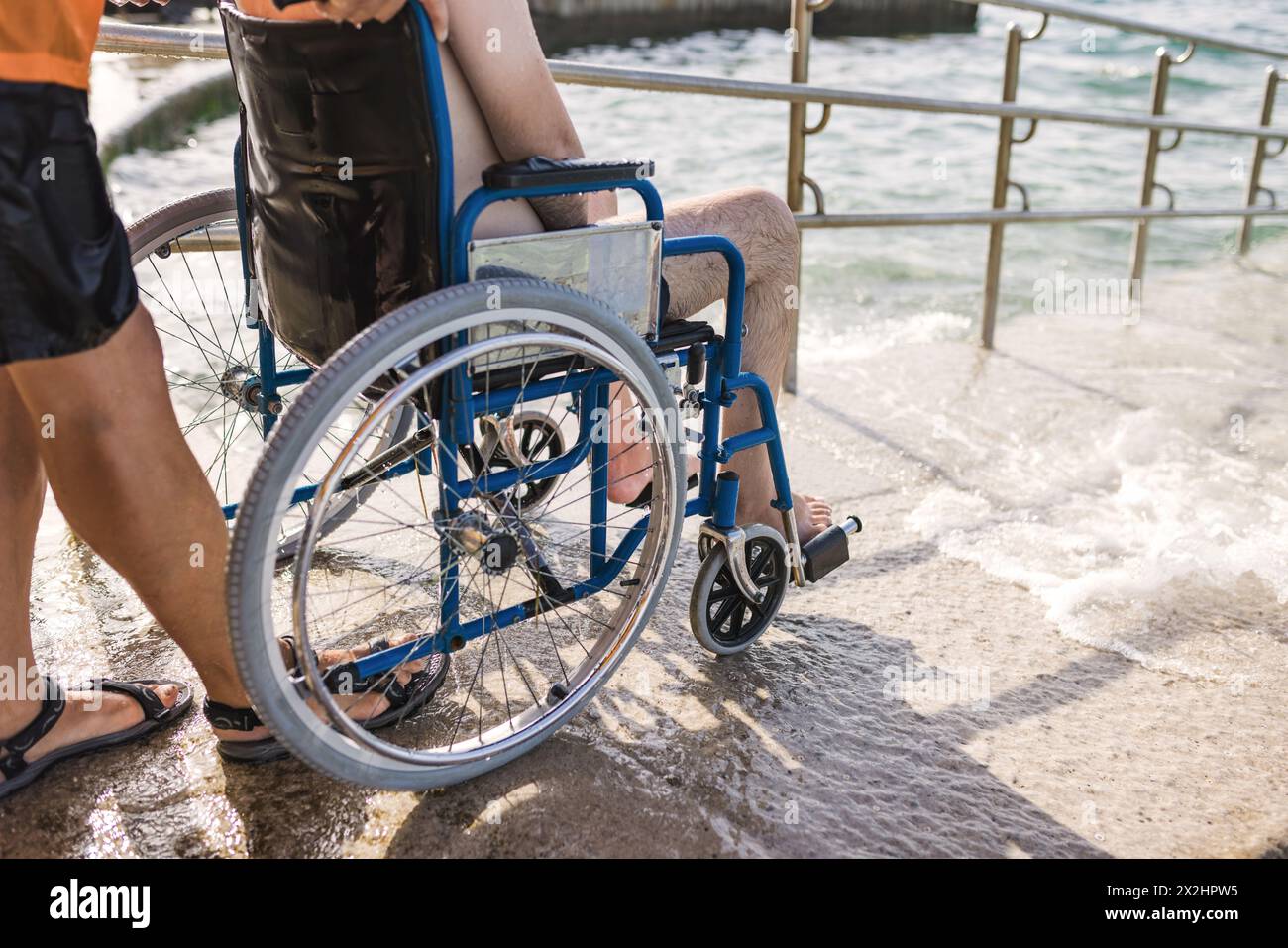 Ein männlicher Assistent hilft einem Mann im Rollstuhl, das Meer auf einer Zufahrtsrampe ins Wasser zu genießen. Barrierefreies Strandkonzept. Stockfoto