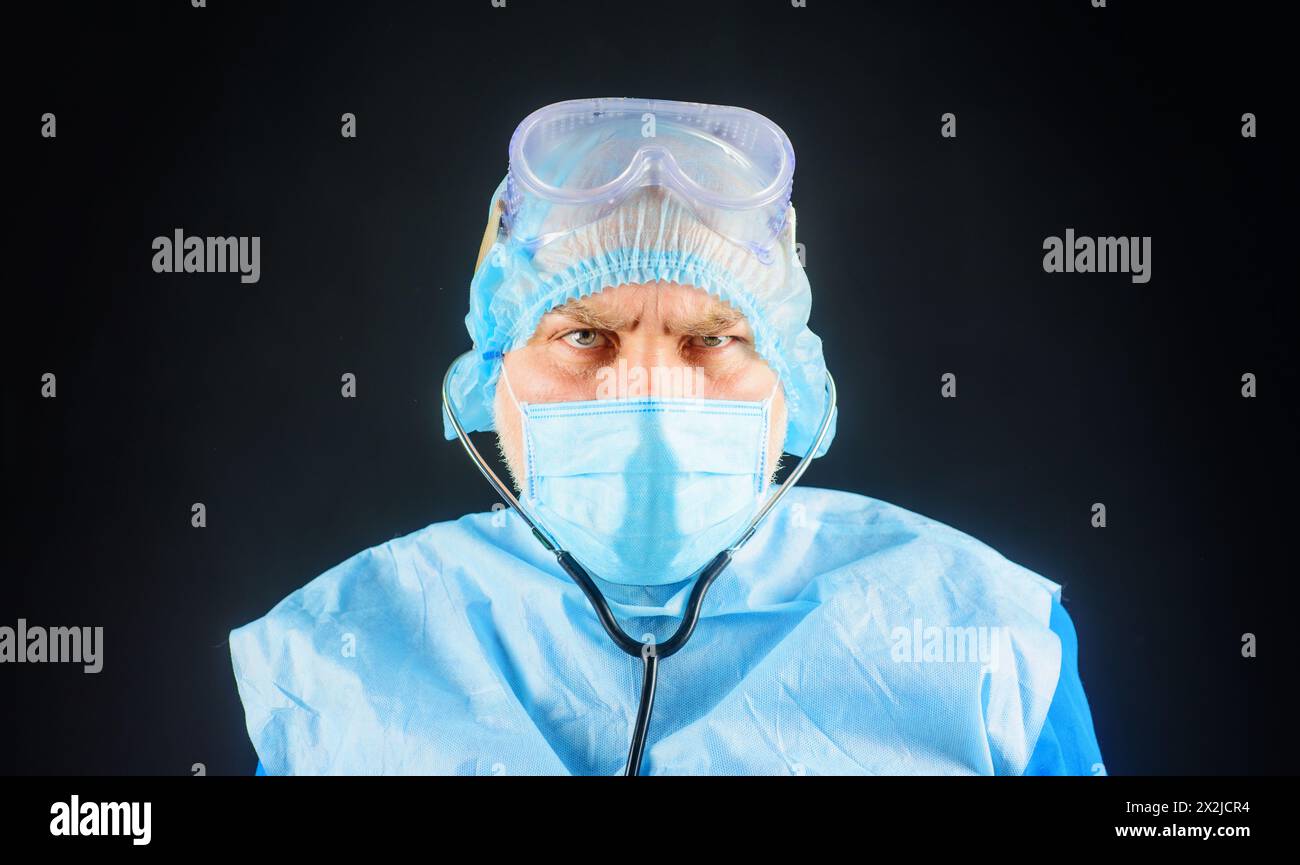 Ernsthafter Arzt in blauer Uniform, Gesichtsmaske, Schutzbrille und Einweg-OP-Kappe. Chirurgenarzt in medizinischer Maske und Peelings mit Stethoskop Stockfoto