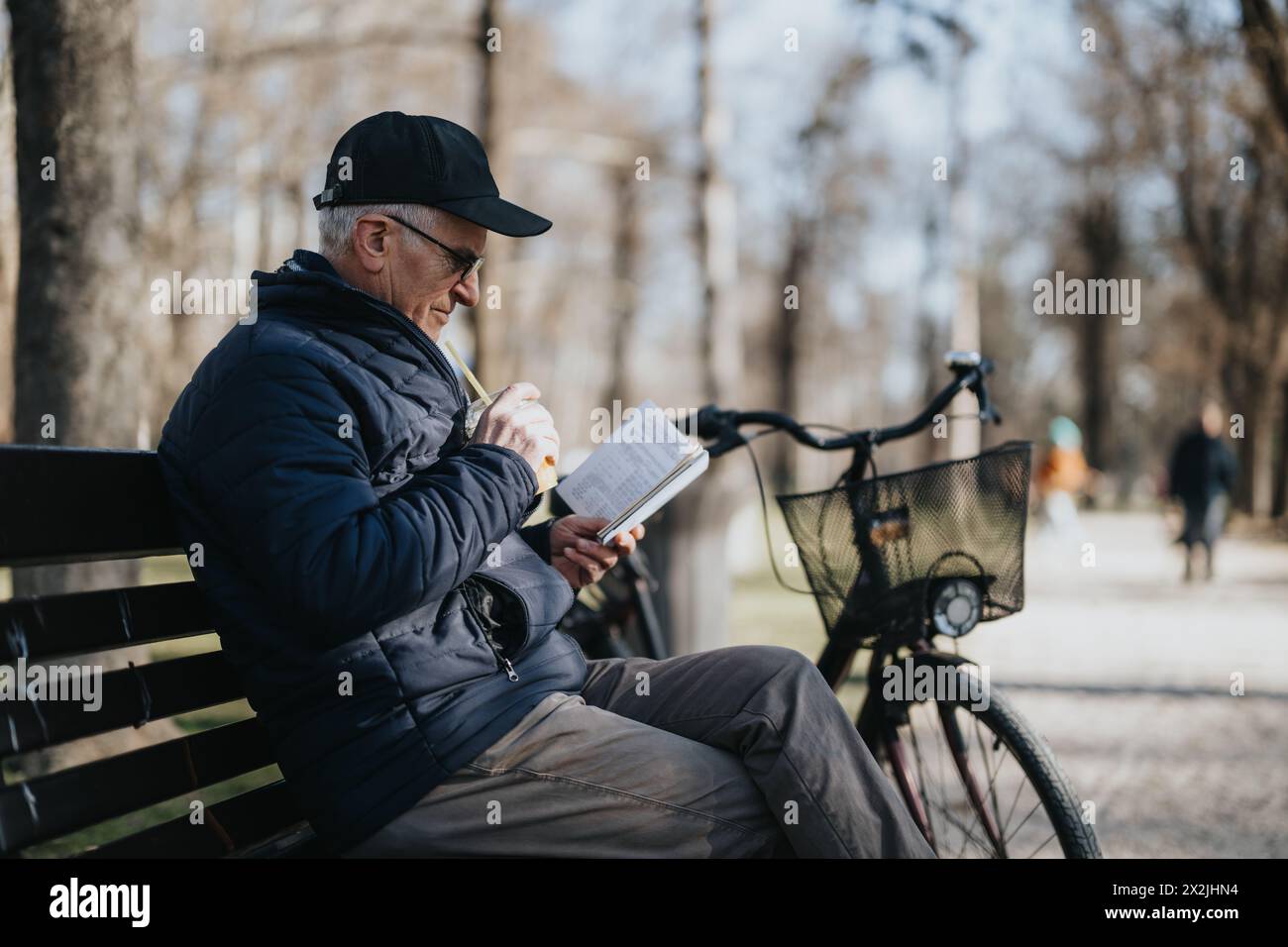 Ein älterer Herr entspannt sich auf einer Parkbank in der Ruhe eines sonnigen Tages, vertieft in das Lesen eines Buches, während sein Fahrrad neben ihm geparkt ist. Stockfoto