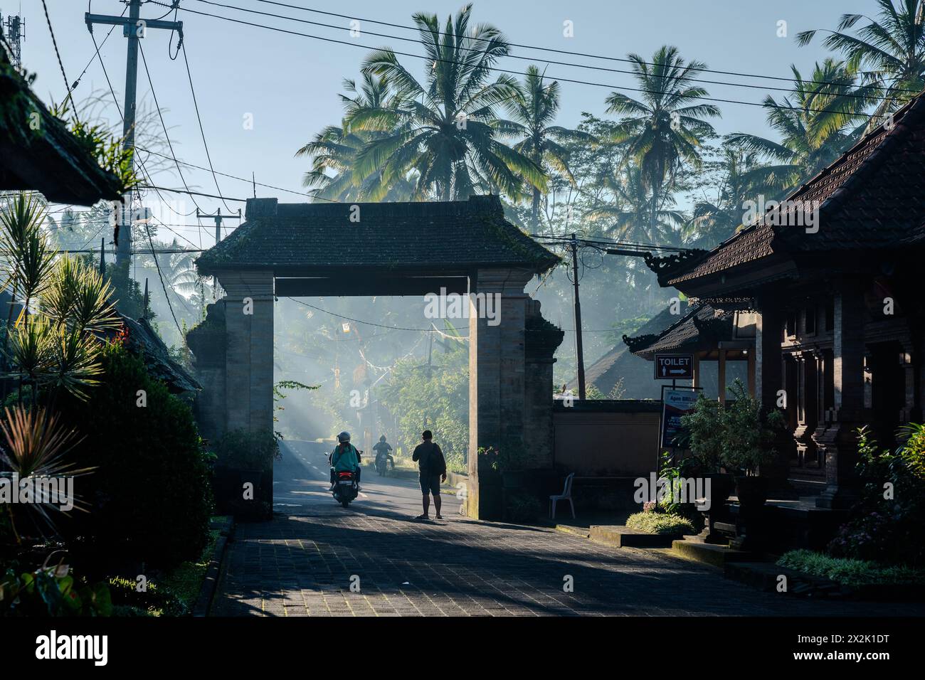 Eine Szene am frühen Morgen in einem tropischen Dorf mit Sonnenlicht, das durch Nebel filtert und Menschen und traditionelle Architektur inmitten von üppigen Bäumen hervorhebt Stockfoto