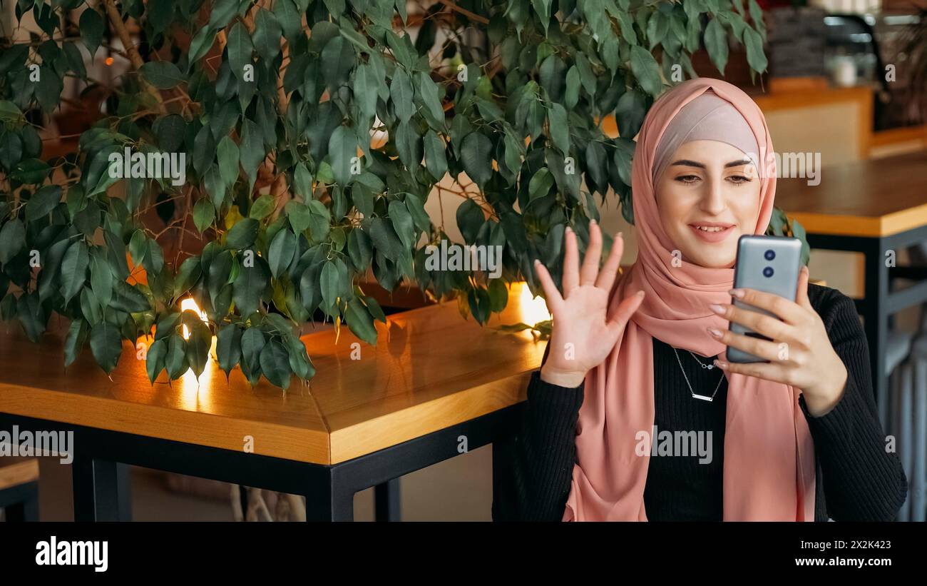 Online-Stream. Videokommunikation. Fröhliche Frau im Hijab winkt Hand zu Followern und grüßt Abonnenten in sozialen Medien im gemütlichen Café. Stockfoto