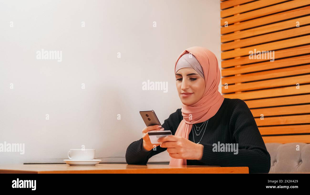 Online-Shopping. Mobiles Banking. Schöne Frau im Hijab, die telefonisch mit Kreditkartenzahlung im Internet im Café-Kopierraum kaufen. Stockfoto