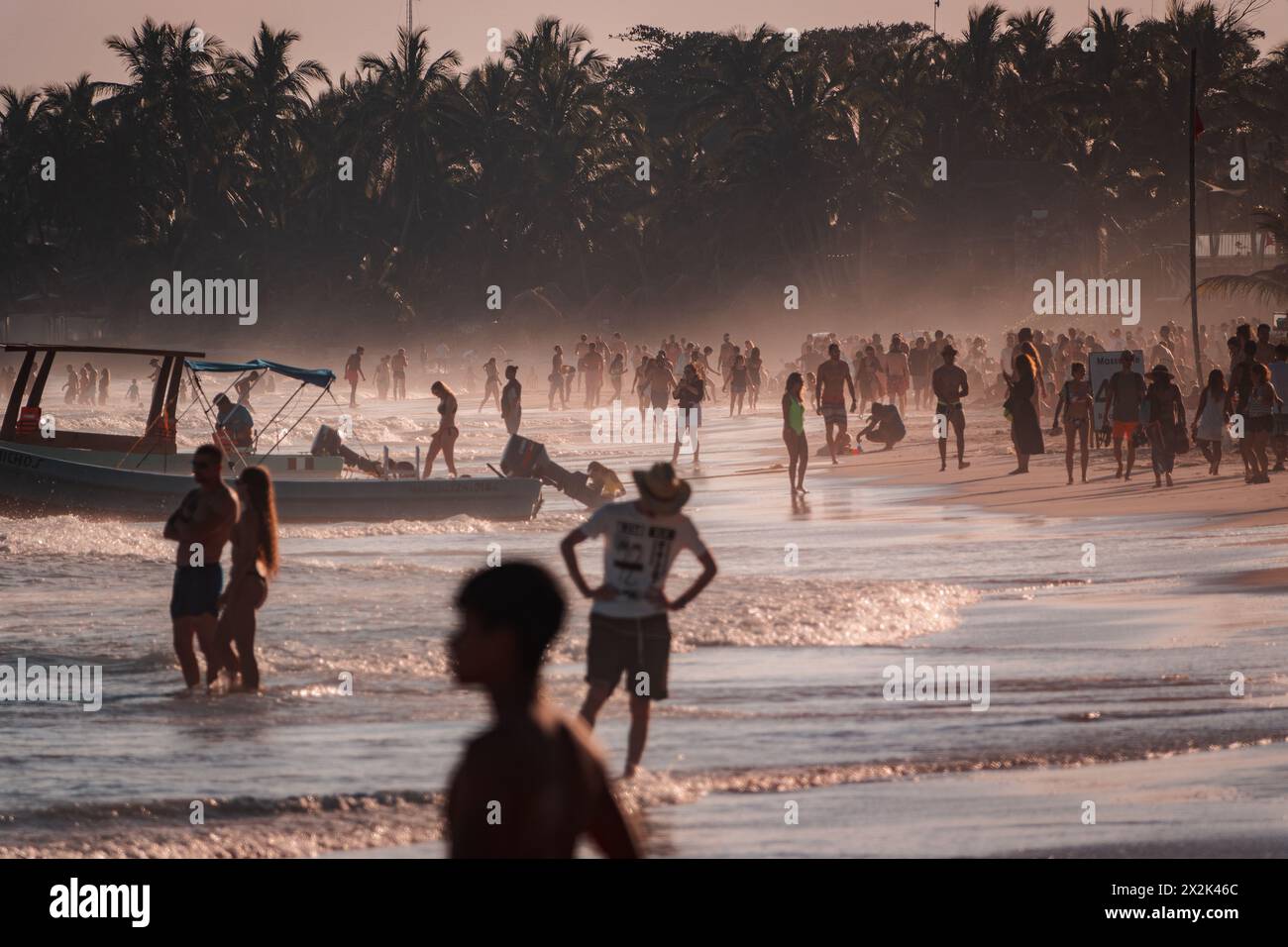 Eine geschäftige Strandszene bei Sonnenuntergang mit Silhouetten zahlreicher Menschen, die den warmen tropischen Sand unter einer Kulisse von Palmen genießen. Stockfoto