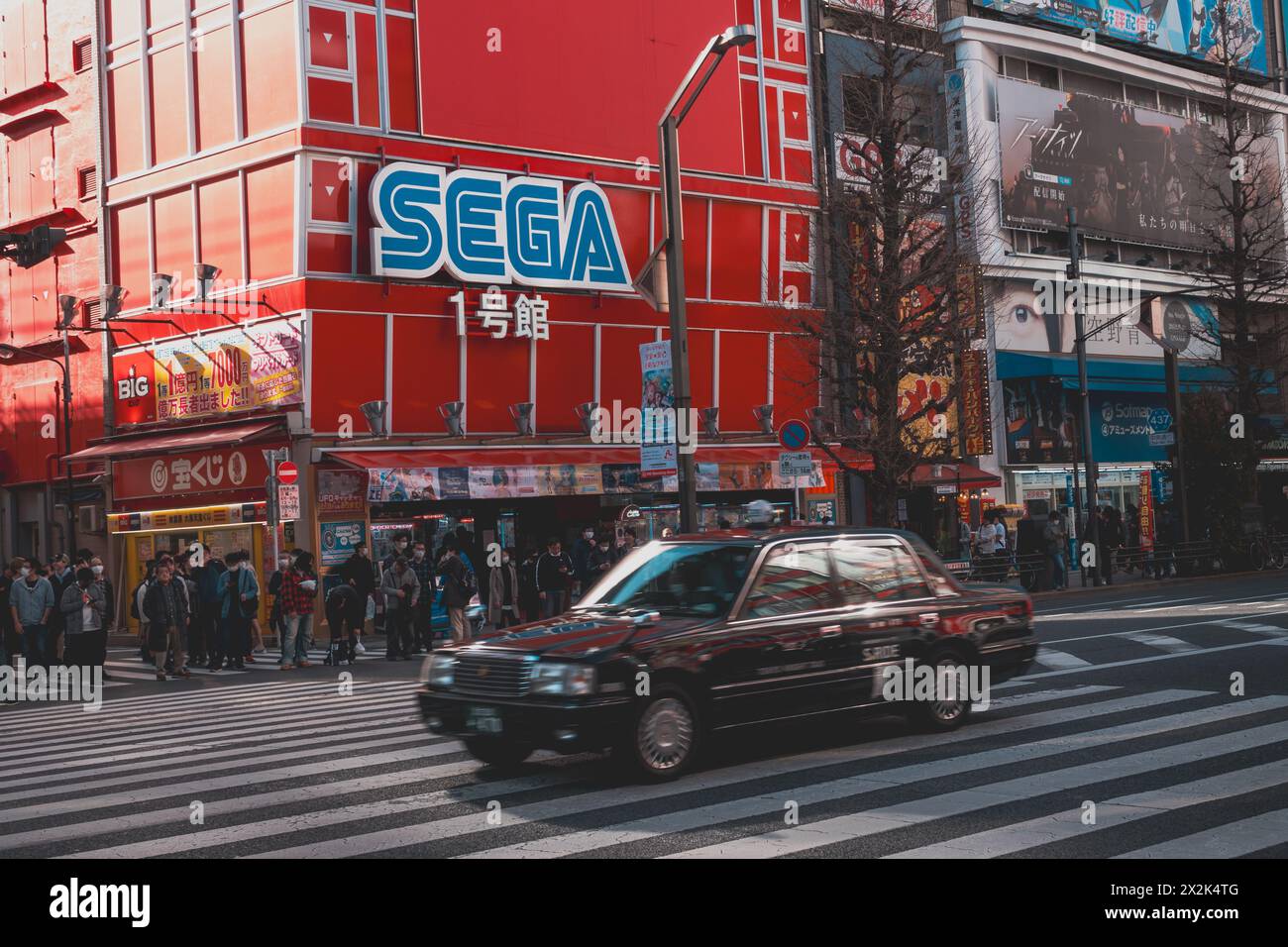 Pulsierende Straßenszene in einer belebten Stadt mit einem prominenten Sega-Gebäude, Fußgängern und einem klassischen schwarzen Taxi, das eine geschäftige Kreuzung überquert. Stockfoto