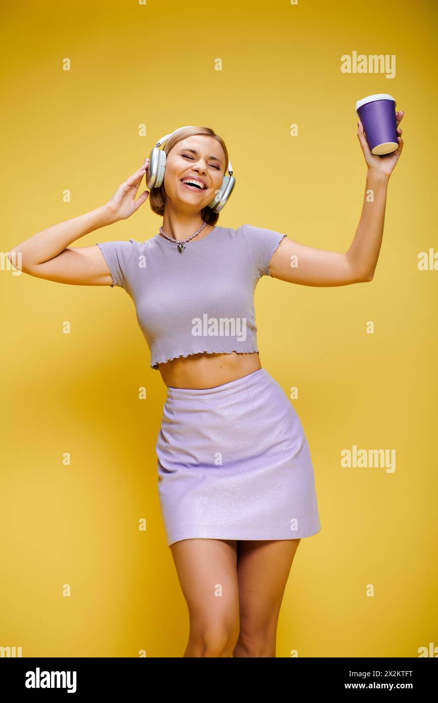 Zufriedene trendige Frau mit Kopfhörern, die Musik und heißen Kaffee genießt, während sie vor gelbem Hintergrund posiert Stockfoto
