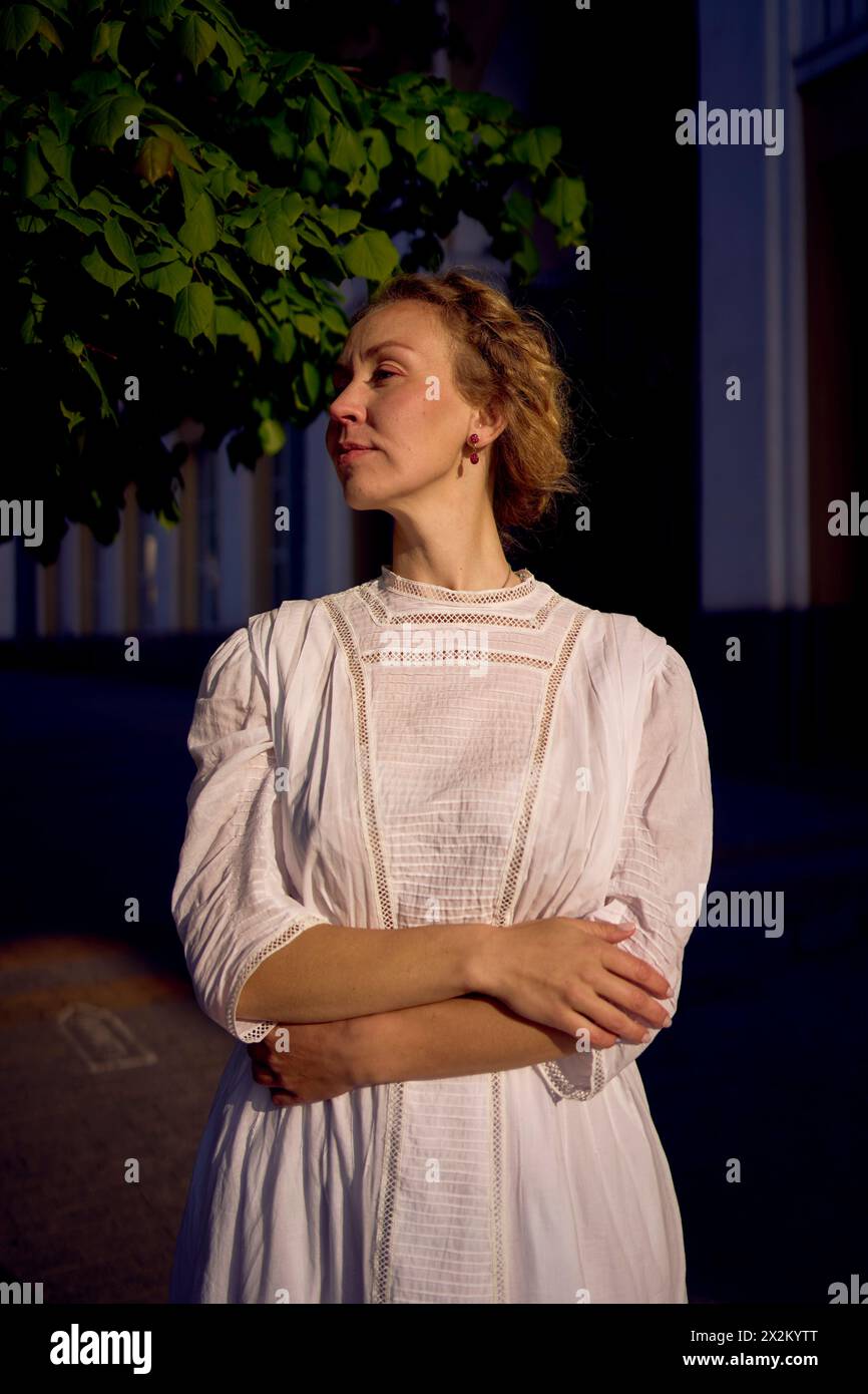 Eine elegante Frau mittleren Alters in einem weißen Vintage-Kleid vor dem Hintergrund historischer Gebäude im Morgenlicht Stockfoto