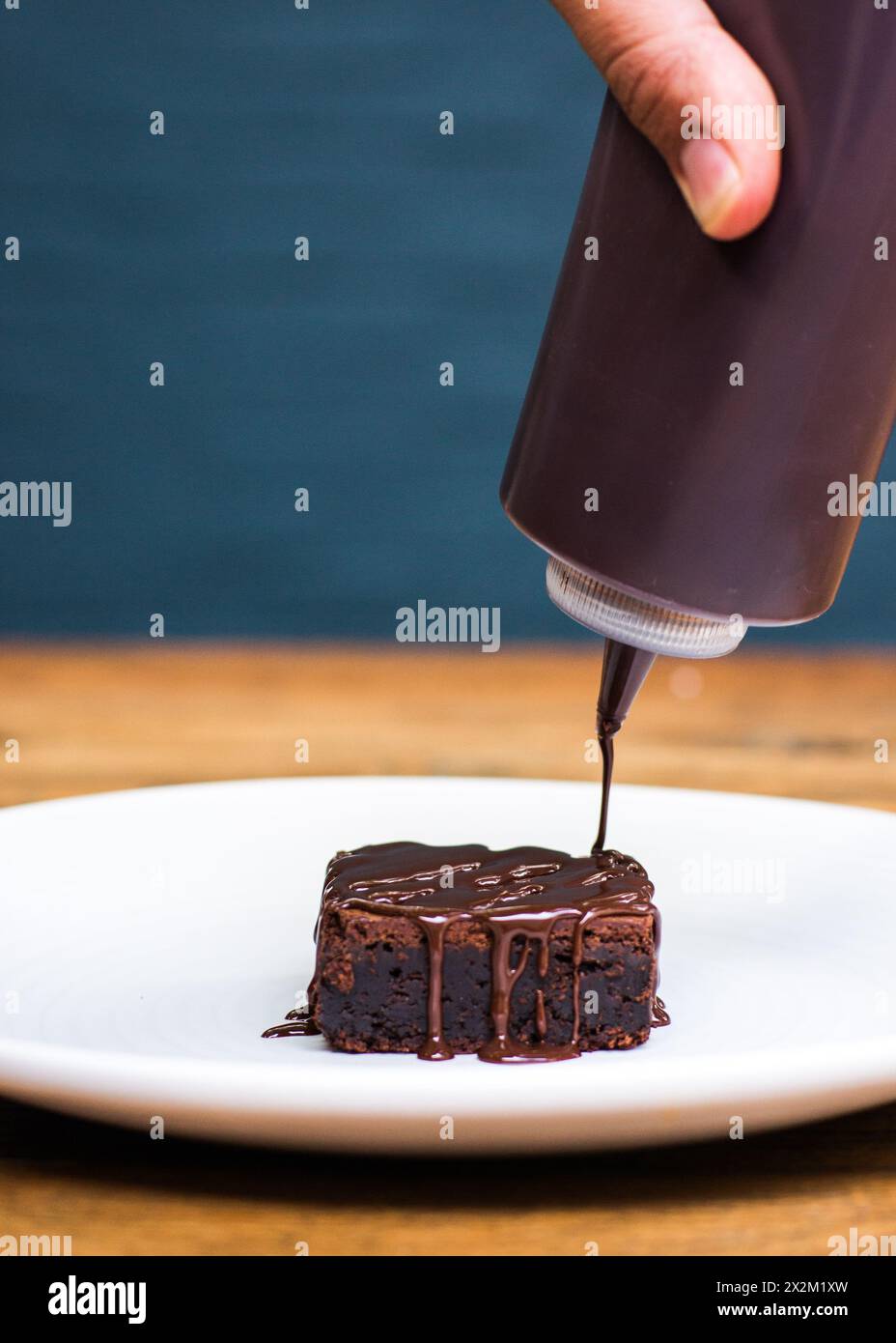 Schokoladensoße wird auf Brownie auf weißem Teller auf hölzernem Restauranttisch gegossen Stockfoto