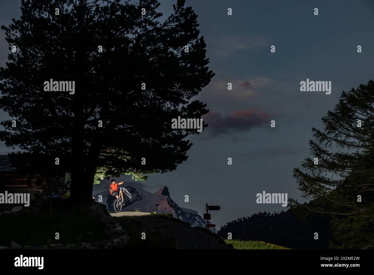 Ein Radfahrer ruht an einem Baum auf einem Bergweg während eines Sommerabends, wobei ein Scheinwerfer die Szene beleuchtet Stockfoto