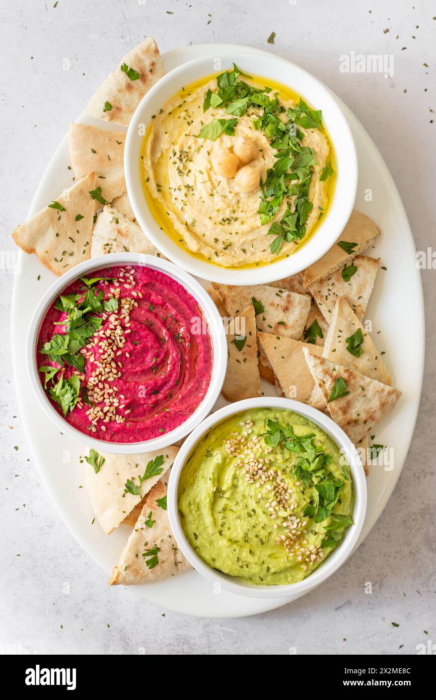 Blick von oben auf die farbenfrohe Auswahl an klassischem Hummus, Rüben und Avocado-Hummus, die jeweils garniert und mit Pita-Brot serviert werden, mit lebendigen, gesunden Dips für Stockfoto