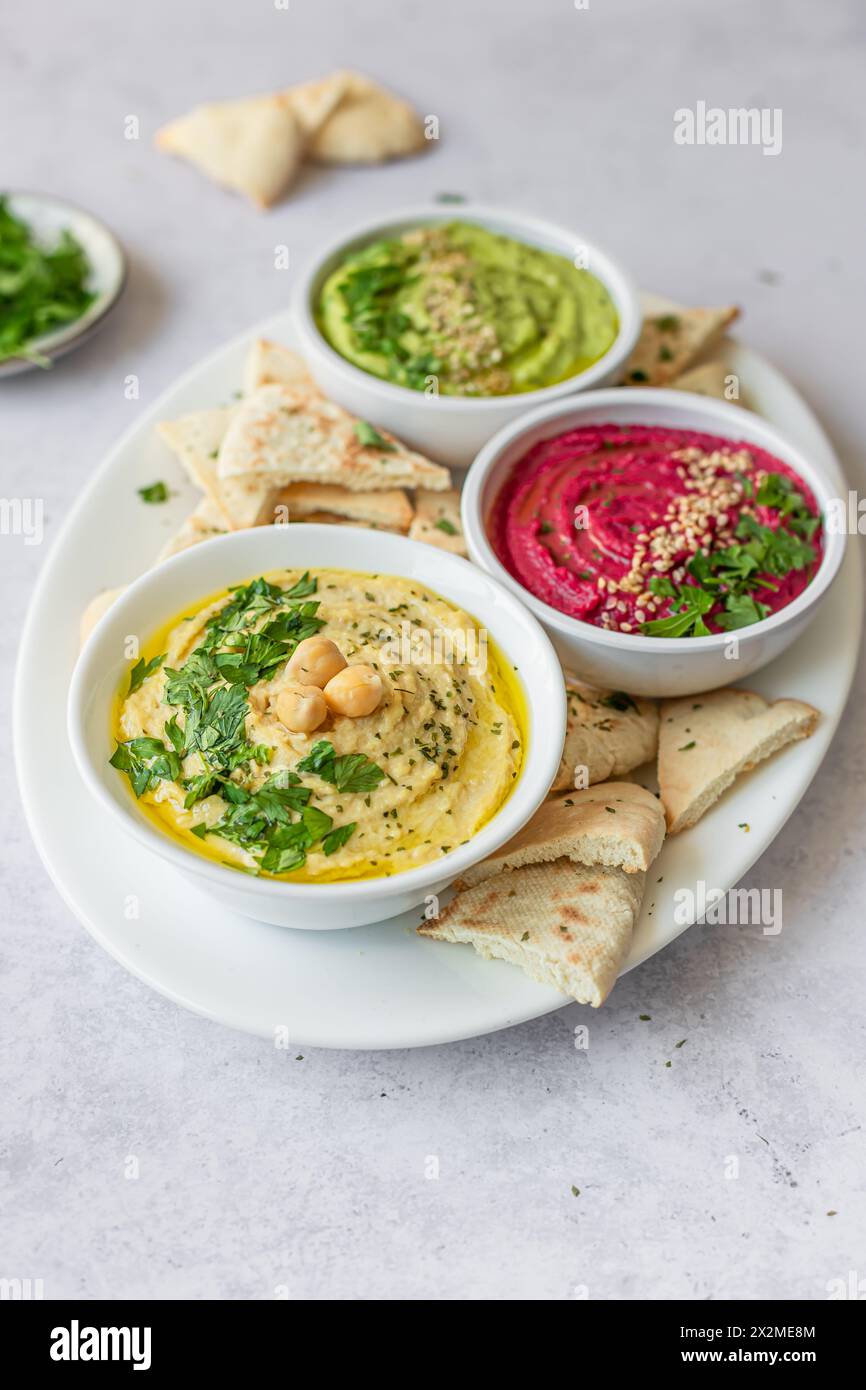 Eine lebhafte Darstellung von drei verschiedenen Hummus-Variationen, Klassik, Rüben und Avocado, jede garniert mit frischen Kräutern und serviert mit Fladenbrot. Stockfoto