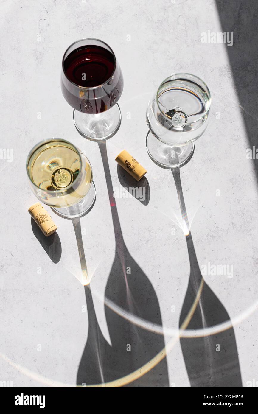 Drei Weingläser, gefüllt mit Rot, weiß und ohne Wein, werfen lange Schatten auf eine strukturierte Oberfläche und zeichnen sich durch ein minimalistisches und elegantes Design aus. Stockfoto