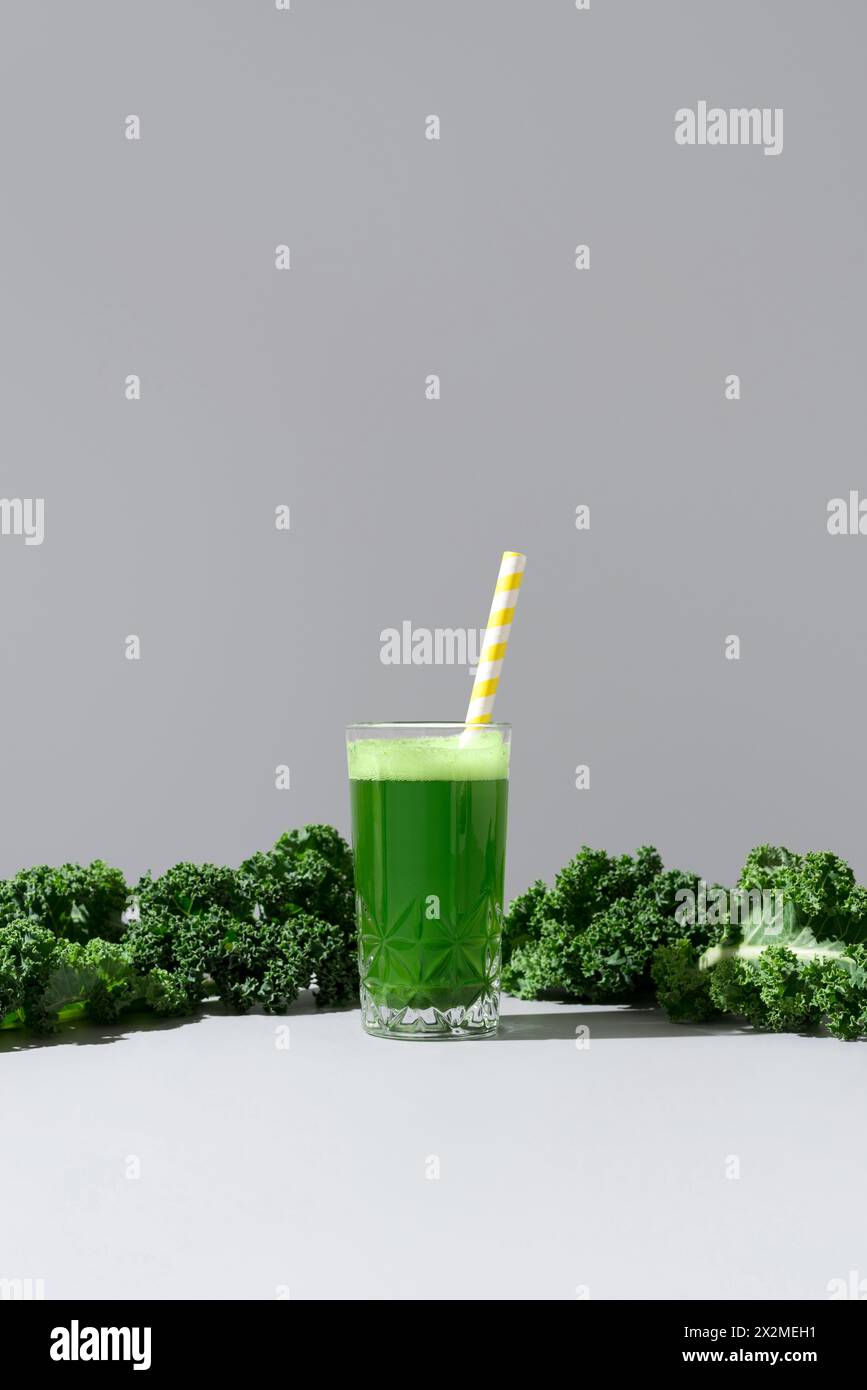 Ein lebhaftes Glas Grünkohl-Saft gepaart mit frischen, lockigen Grünkohlblättern auf neutralem Hintergrund, mit einem gelb gestreiften Strohhalm für eine frische Ästheti Stockfoto