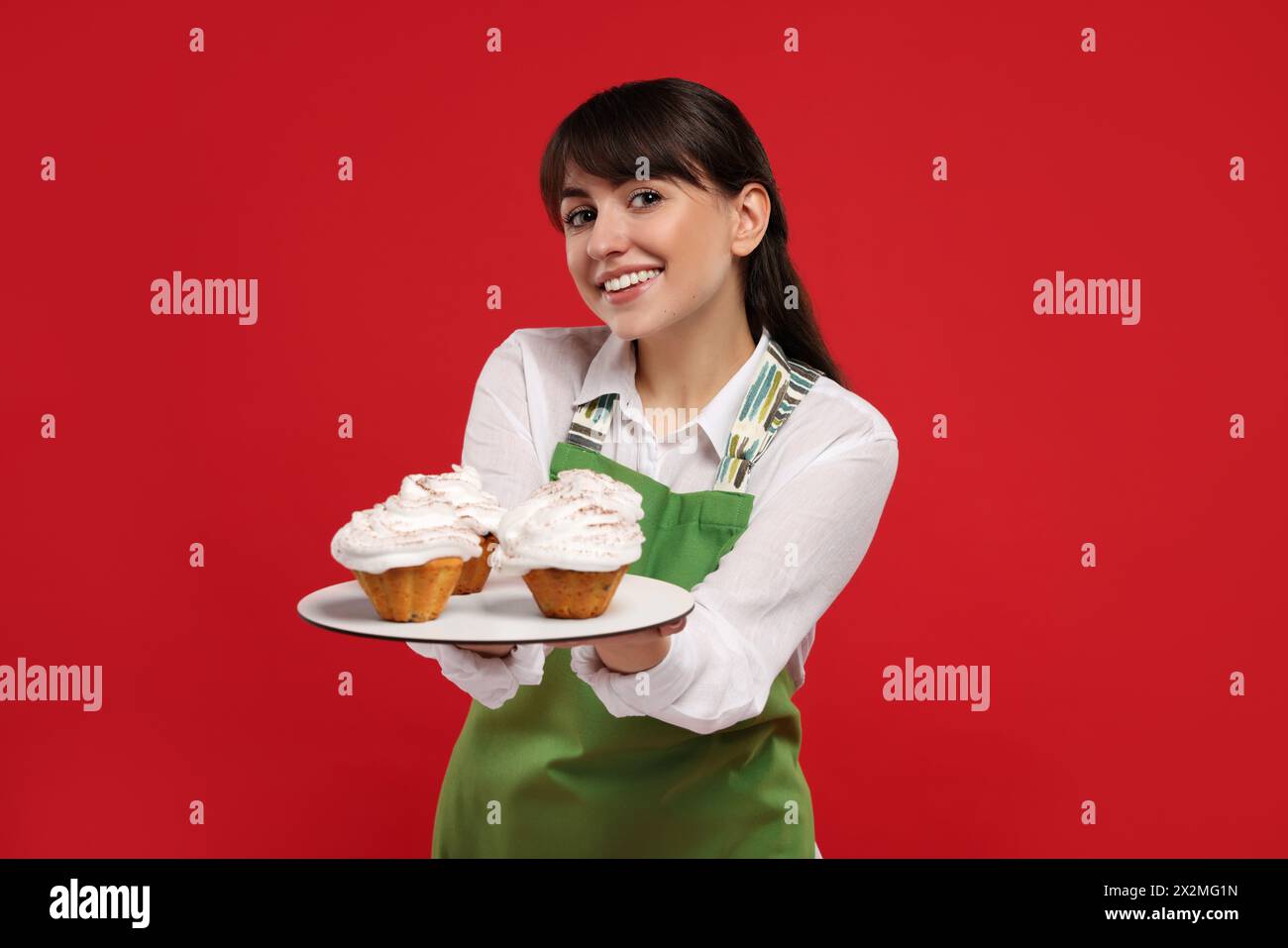 Glücklicher professioneller Konditor in Schürze mit köstlichen Cupcakes auf rotem Hintergrund Stockfoto