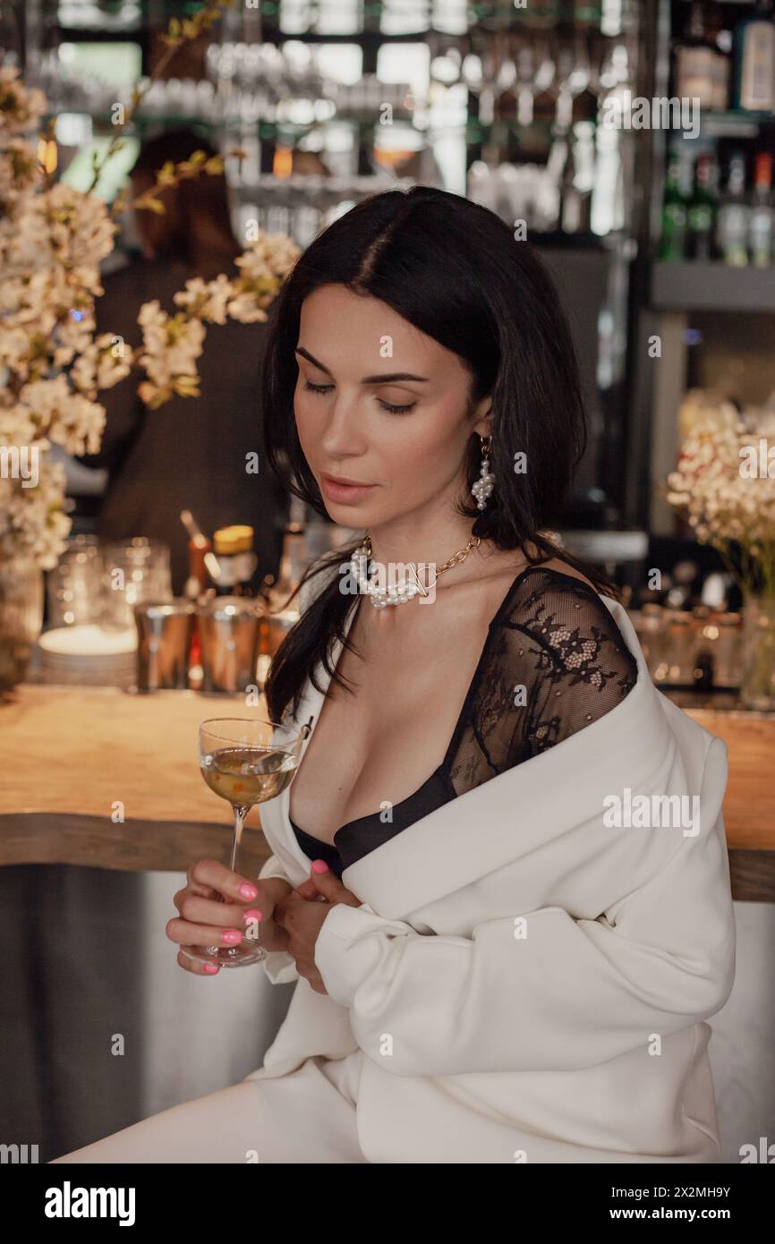 Eine Frau sitzt in einer Bar, hält ein Glas Wein, taucht in ihre Gedanken ein und genießt ihre Zeit am Veranstaltungsort. Stockfoto