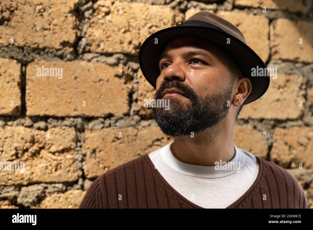 Latino-Mann (39) in braunem Hut und Pullover, schaut auf sein Handy, während er mit seinen Augen eine nicht konforme Geste macht. Lifestyle-Konzept. Stockfoto