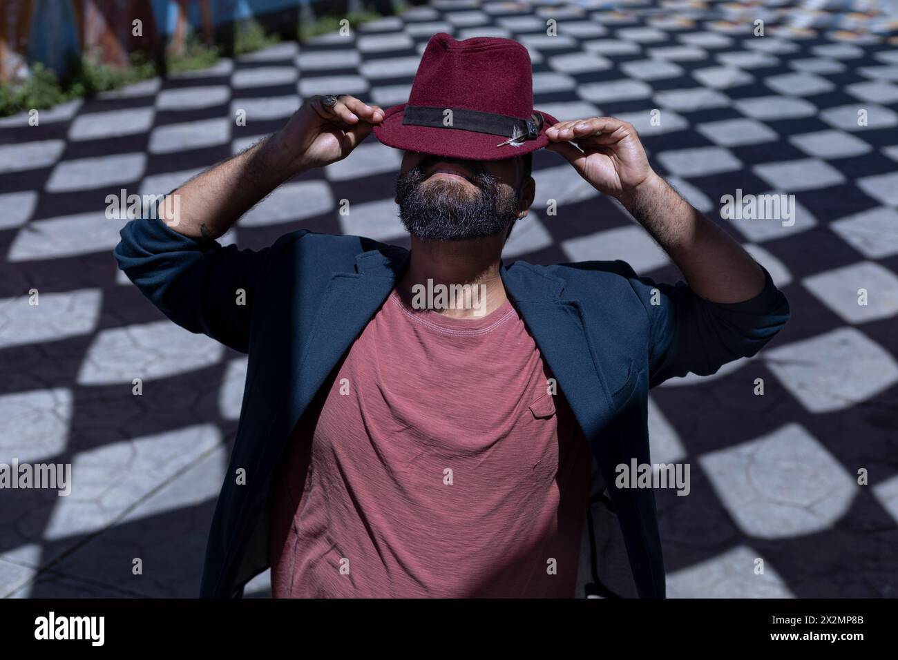 Porträt eines lateinamerikanischen Künstlers mit Bart, Hut und Jacke. Er trägt seinen Hut auf komische Art und Weise und versteckt sein Gesicht. Hintergrund von Schatten mit r Stockfoto