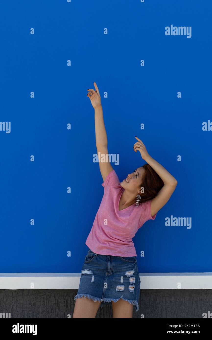 Mittellange Aufnahme einer jungen Frau (22) lateinamerikanische Rothaarige, die mit beiden Armen nach oben blickt und zeigt. Blauer Wandhintergrund. Vertriebskonzept Stockfoto