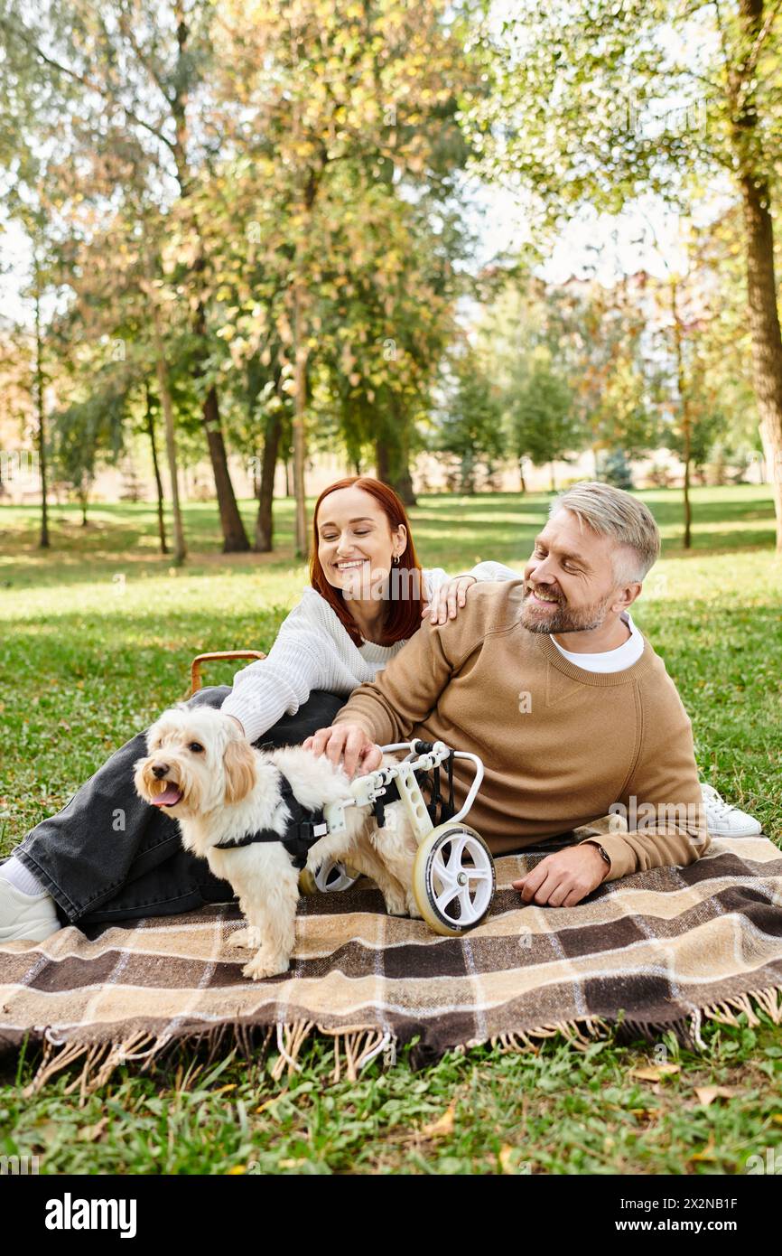 Ein Mann und eine Frau in lässiger Kleidung sitzen mit ihrem Hund in einer friedlichen Parklandschaft auf einer Decke. Stockfoto
