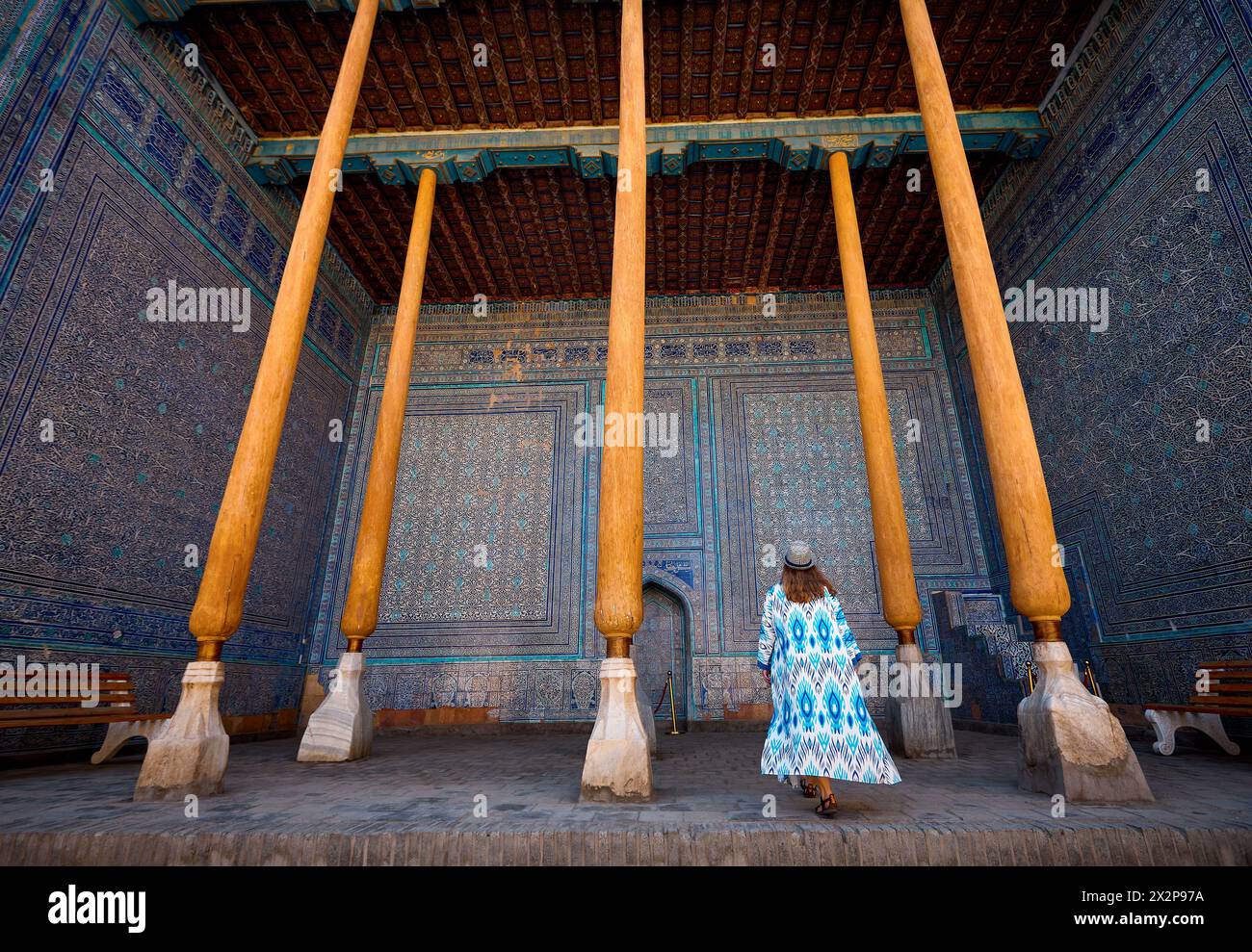 Frau Touristin in ethnischer Kleidung im Innenhof des blauen Mosaikpalastes in Tash Hauli mit Holzsäule in der antiken Stadt Chiwa in Usbekistan. Stockfoto