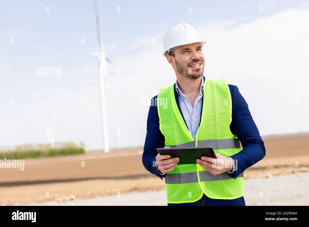 Ein fröhlicher Ingenieur in Sicherheitsausrüstung überprüft die Daten auf einem Tablet, bevor Windmühlen unter klarem Himmel aufsteigen. Stockfoto