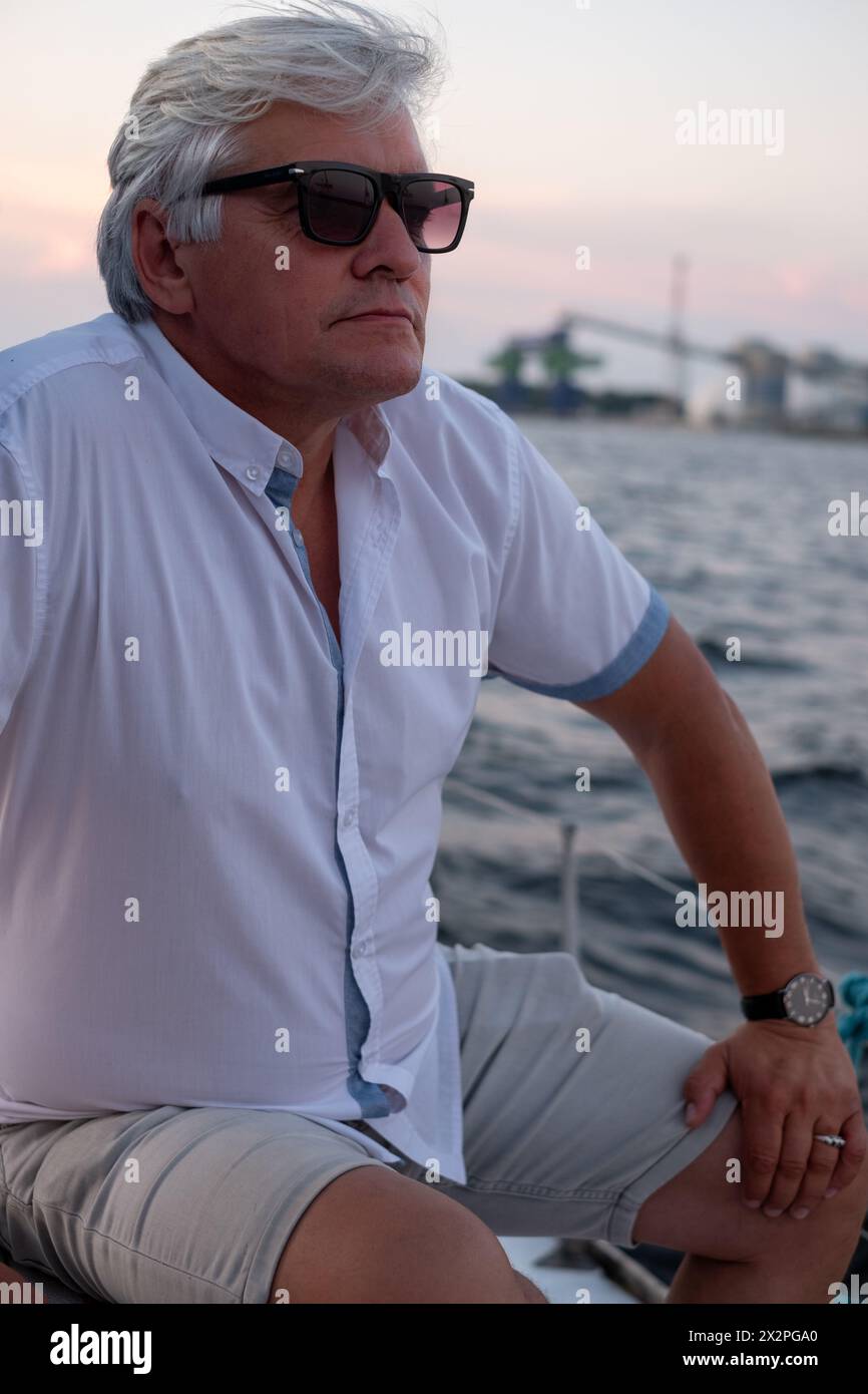 Silberhaariger Mann in Sonnenbrille, nachdenklich auf das Meer von einem Boot in der Abenddämmerung aus blickt und eine Stimmung der Reflexion und Einsamkeit einfängt. Hochwertige Fotos Stockfoto