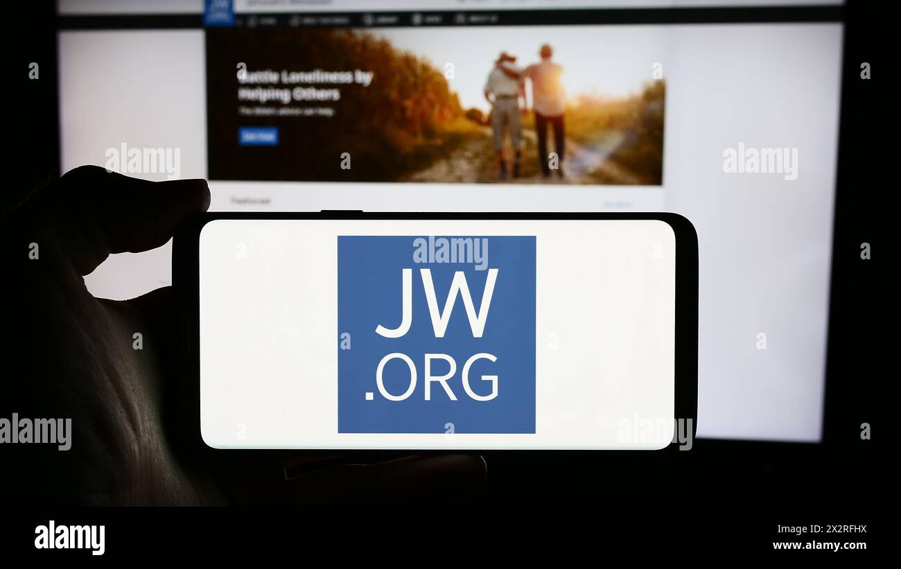 Person mit Handy mit Logo der religiösen Organisation Zeugen Jehovas vor der Webseite. Schwerpunkt auf dem Display des Telefons. Stockfoto