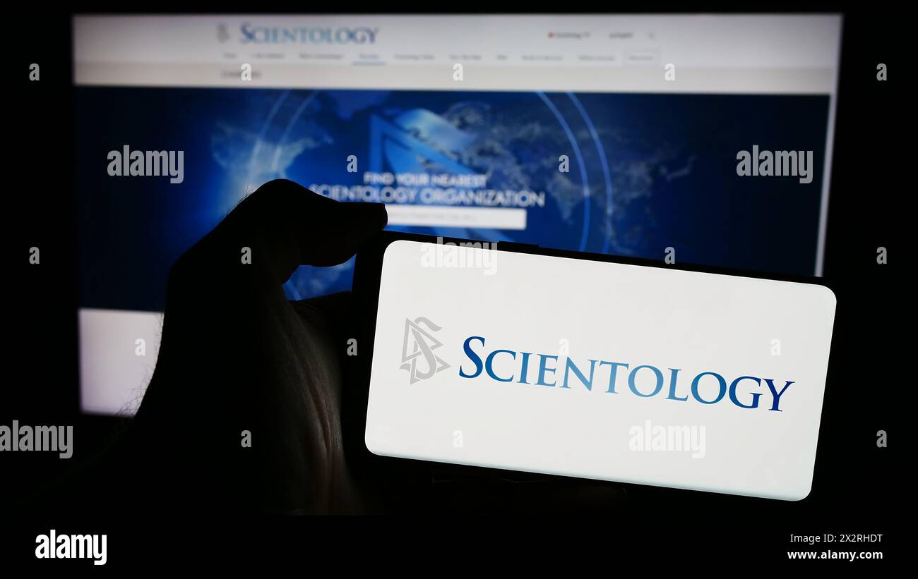 Person, die ein Mobiltelefon mit dem Logo der religiösen Organisation Scientology vor der Webseite hält. Schwerpunkt auf dem Display des Telefons. Stockfoto