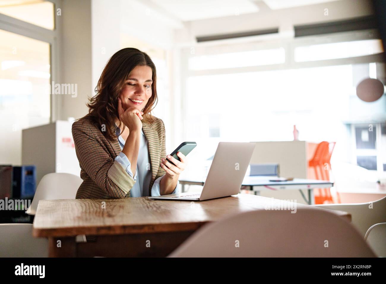 Lächelnde Geschäftsfrau, die ihr Mobiltelefon am Laptop-Schreibtisch im Büro benutzt Stockfoto