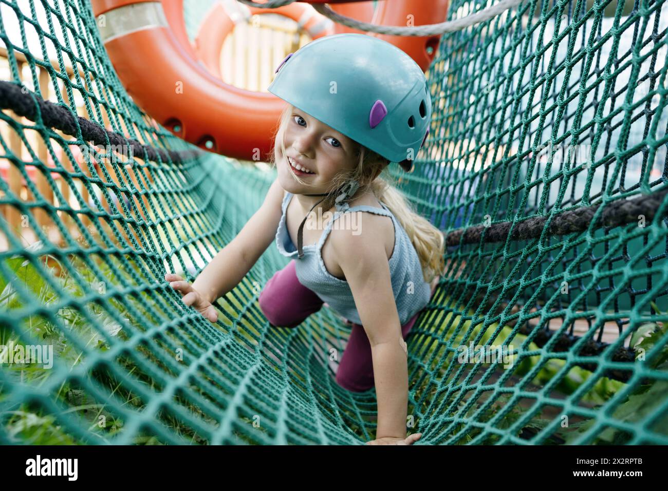 Glückliches Mädchen, das Helm trägt und im Park im Netz krabbelt Stockfoto
