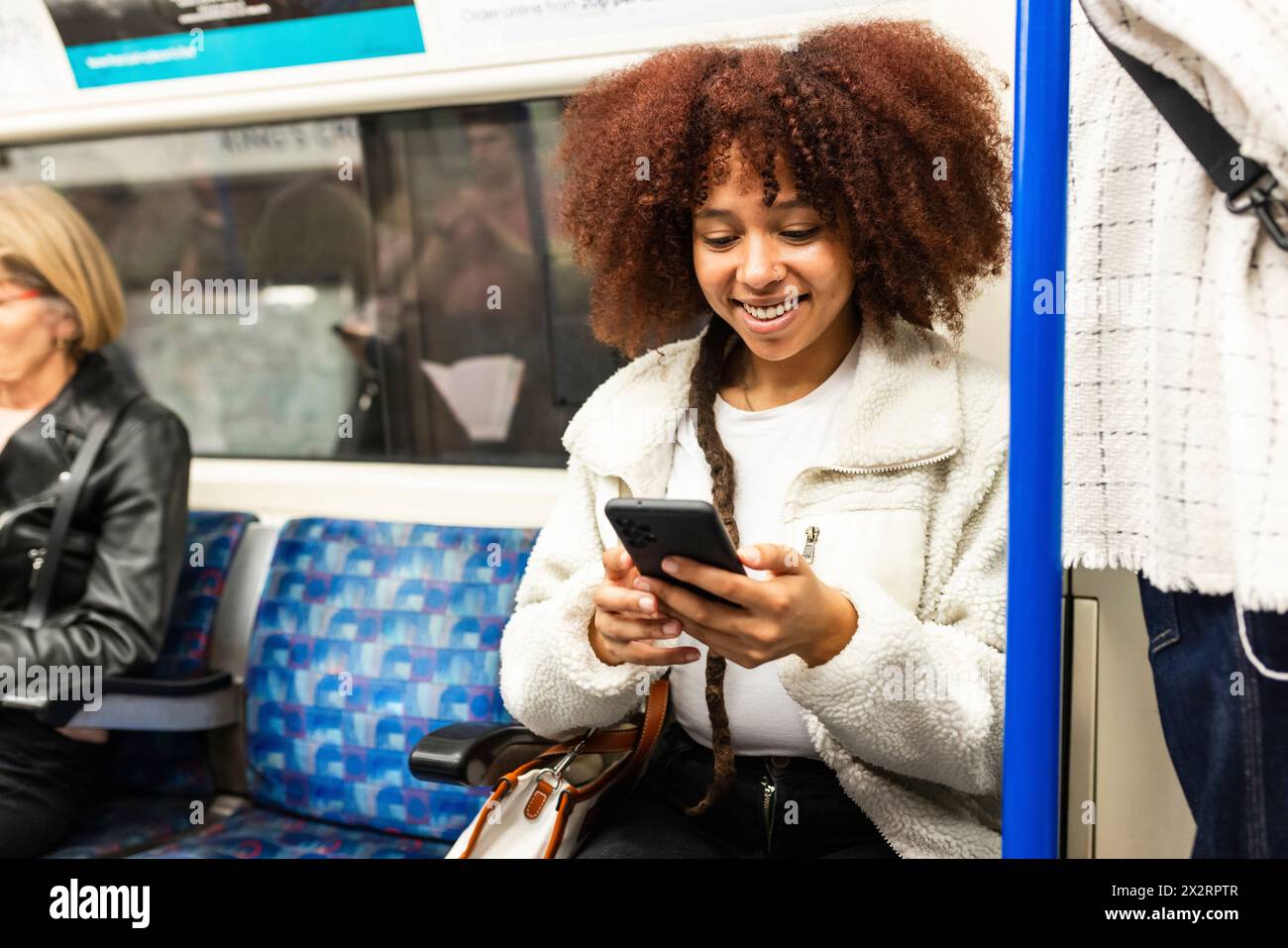 Lächelnde junge Frau, die in der U-Bahn sitzt und ihr Smartphone benutzt Stockfoto