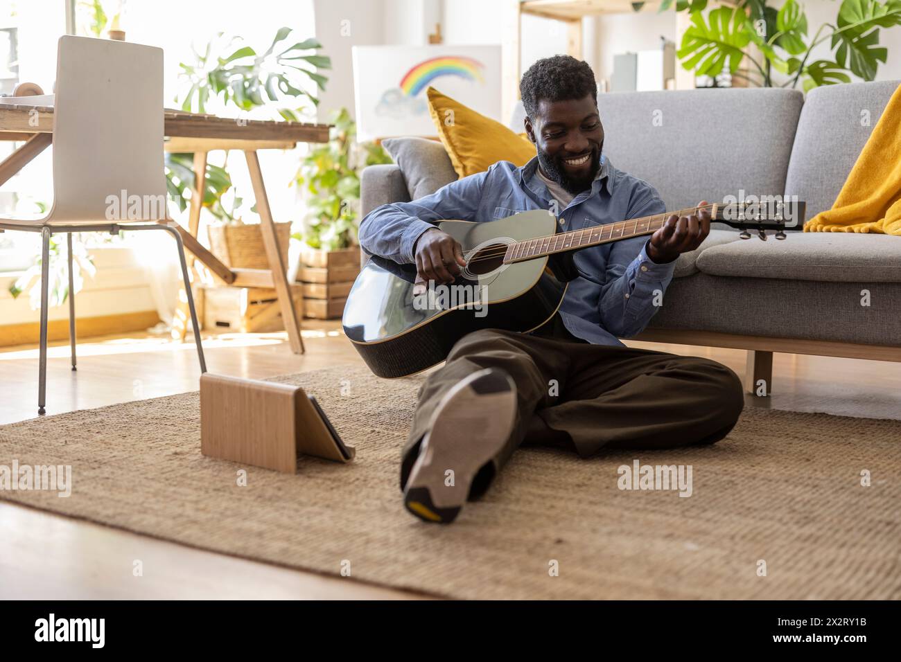 Glückliche, nicht binäre Person, die zu Hause in der Nähe eines Tablet-PCs Gitarre spielt Stockfoto