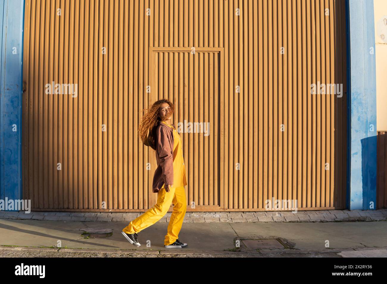 Lächelnde junge Frau in Jacke, die auf einem Fußweg nahe der orangefarbenen Metallwand läuft Stockfoto