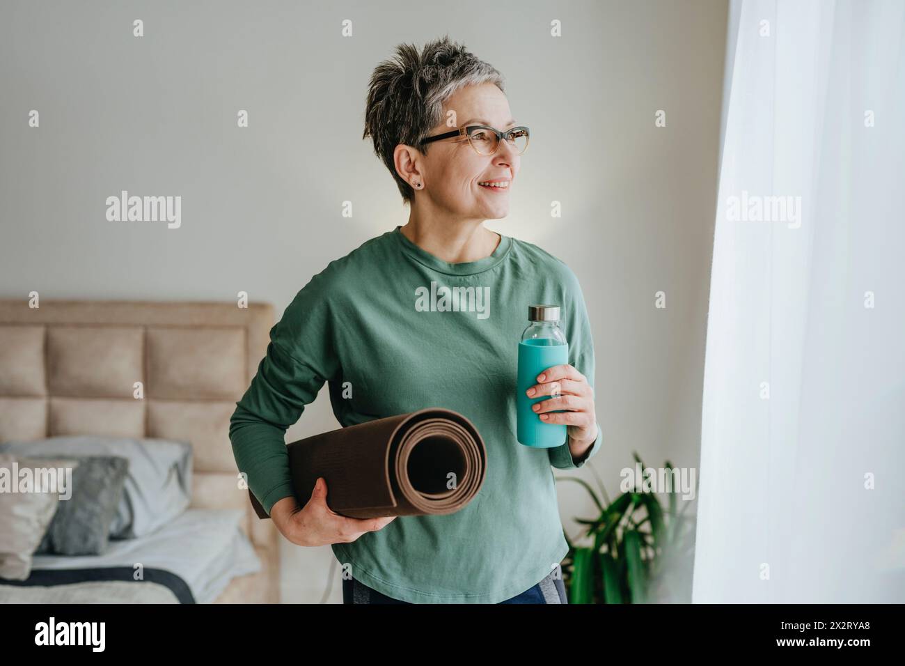 Lächelnde Frau, die zu Hause Yogamatte und Wasserflasche hält Stockfoto