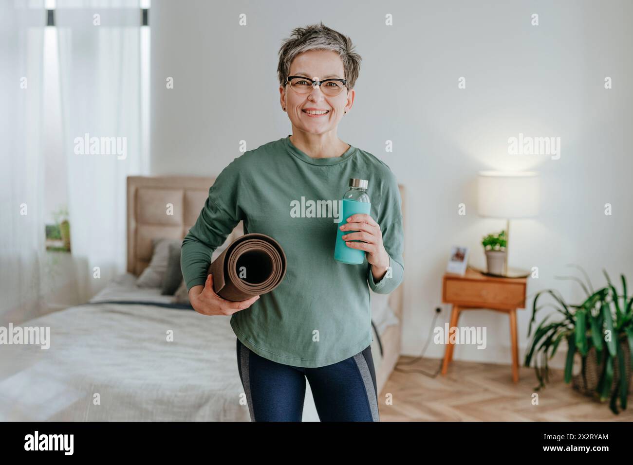 Lächelnde reife Frau, die zu Hause Wasserflasche und Yogamatte hält Stockfoto