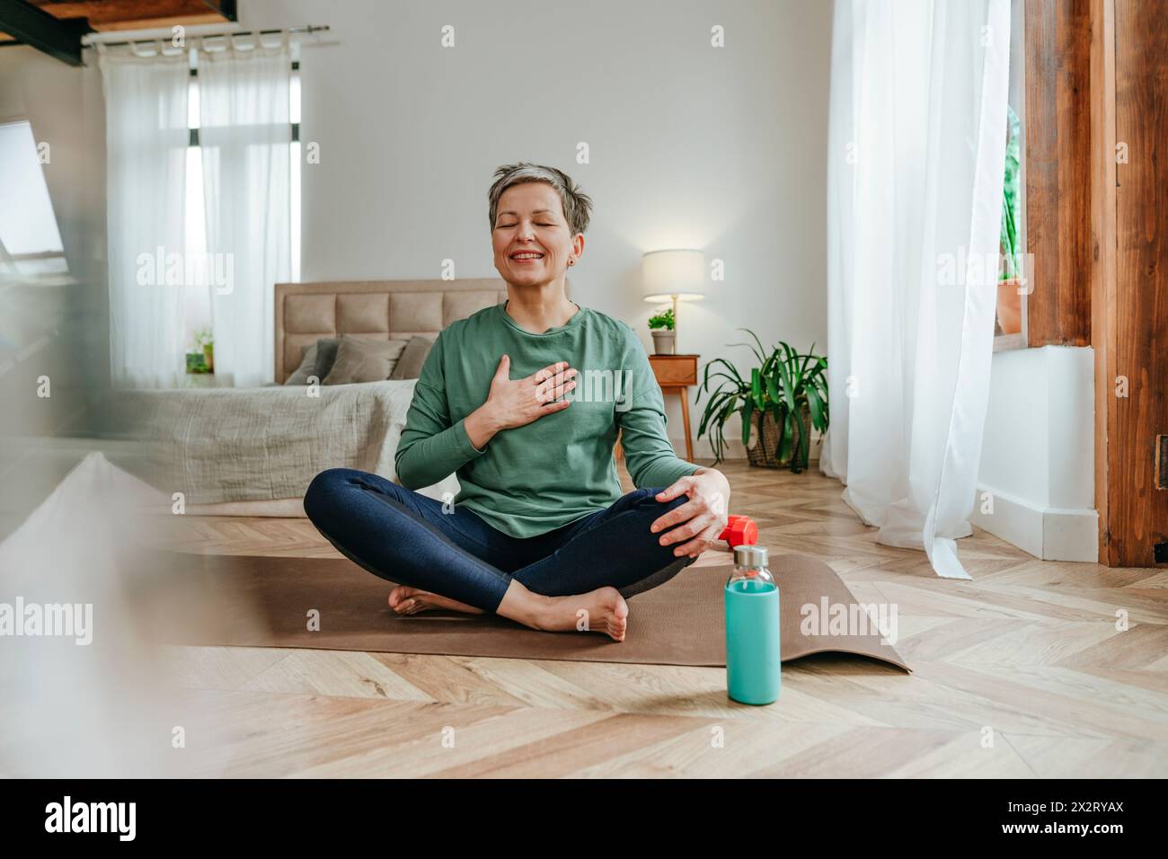 Lächelnde Frau mit Hand auf der Brust sitzend auf Yogamatte zu Hause Stockfoto