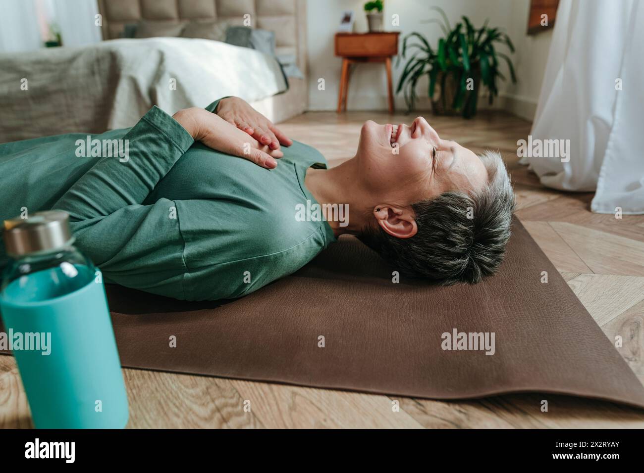 Lächelnde Frau mit Händen auf der Brust, die zu Hause auf Yogamatte liegt Stockfoto