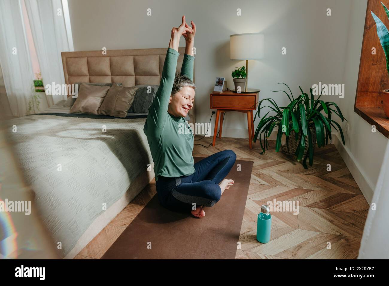 Lächelnde Frau, die sich auf Yogamatte in der Nähe des Bettes zu Hause ausdehnt Stockfoto