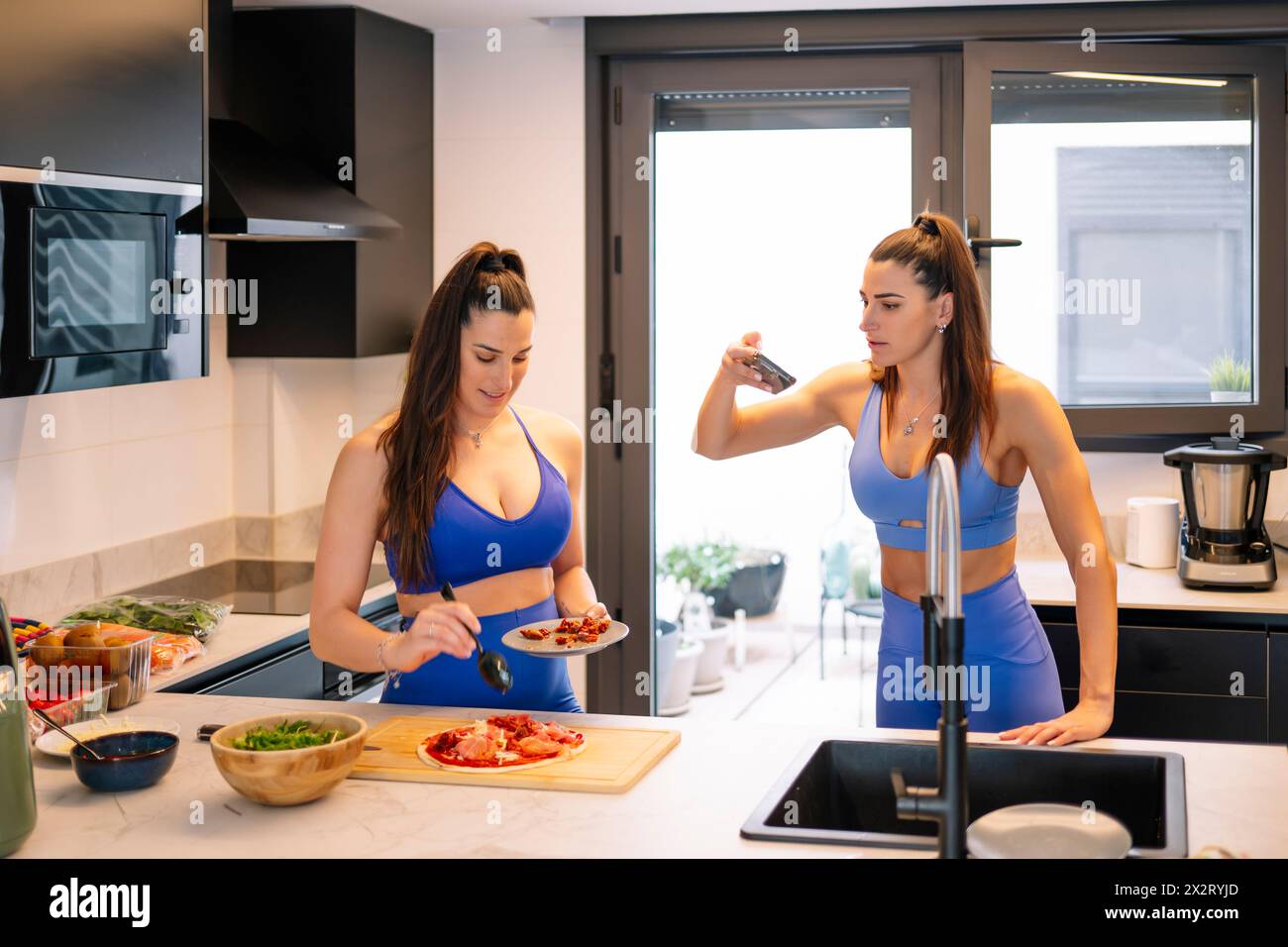Frau, die Fotos von Schwester macht, die zu Hause Essen in der Küche zubereitet Stockfoto