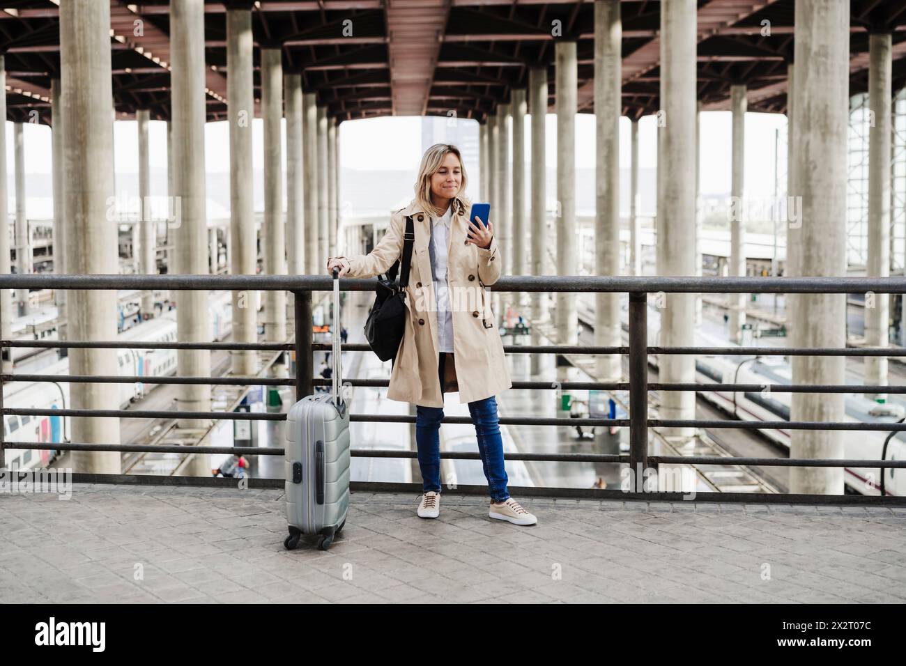 Frau, die ihr Smartphone benutzt und am Bahnhof mit Gepäck steht Stockfoto