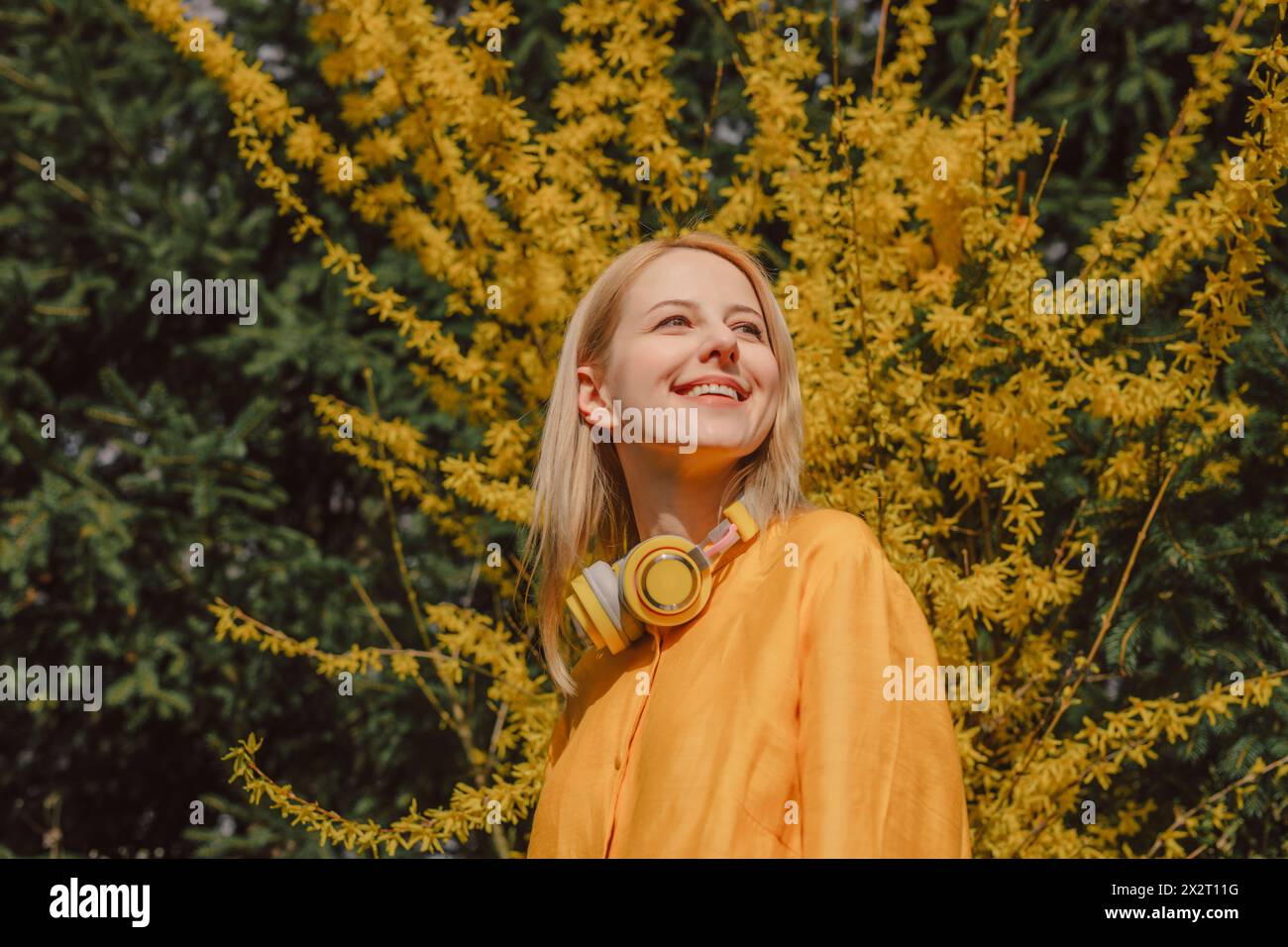 Lächelnde Frau mit kabellosen Kopfhörern, die neben dem gelben Blütenbaum steht Stockfoto