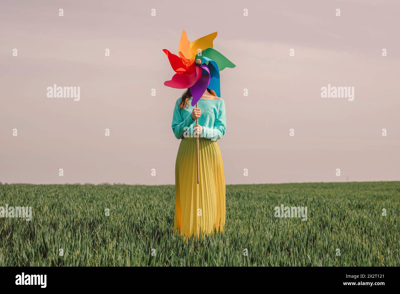 Junge Frau, die ein mehrfarbiges Nadelrad-Spielzeug hält und im grünen Weizenfeld unter dem Himmel steht Stockfoto