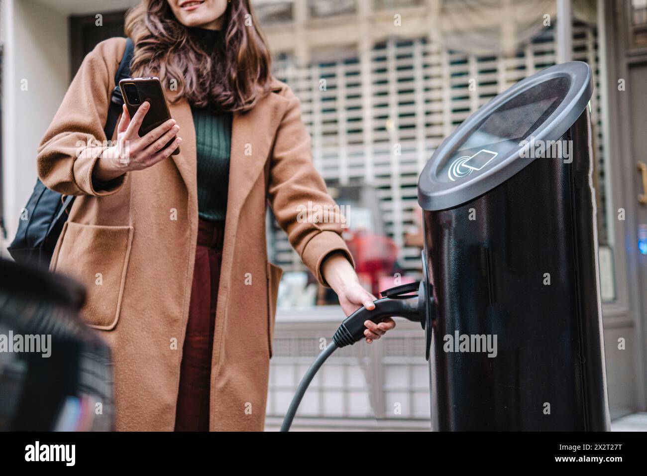 Geschäftsfrau mit Smartphone, das den Elektrostecker an der Fahrzeugladestation hält Stockfoto